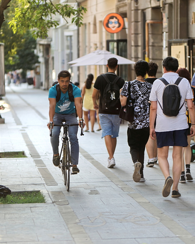 8 - A person riding a bicycle on a pedestrian walkway with a painted bike symbol on the pavement, surrounded by walking pedestrians